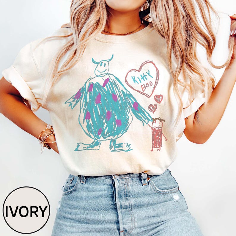 May include: Ivory-colored t-shirt featuring a whimsical, hand-drawn design. The design includes a teal monster with purple spots, a heart with the words "Kitty Boo," and two small hearts. The shirt is a relaxed fit.