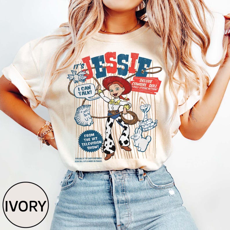 May include: Ivory t-shirt with a retro graphic of Jessie, the cowgirl doll, and the text "It's Jessie." The design includes the phrases "I can talk!" and "Deluxe Cowgirl Doll." The shirt has a vintage style.