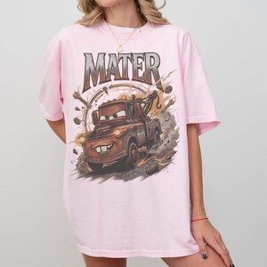Puede incluir: Camiseta rosa claro con una ilustraci&oacute;n de Mater de la pel&iacute;cula Cars. La palabra "MATER" est&aacute; impresa encima de la imagen en una fuente met&aacute;lica. El gr&aacute;fico muestra una gr&uacute;a marr&oacute;n con una cara sonriente, sobre un fondo de rayos y humo.