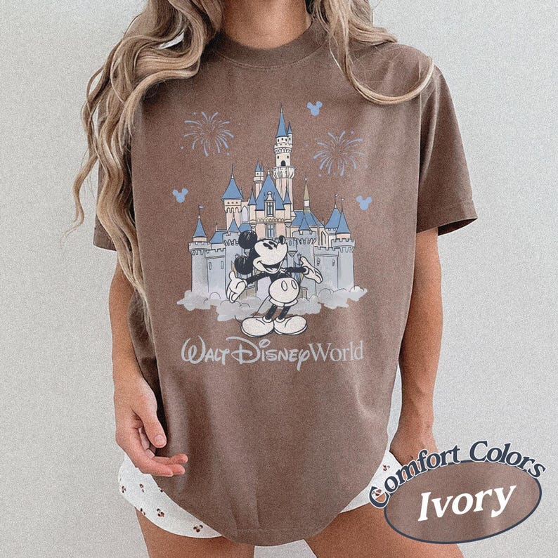 May include: A brown t-shirt with a graphic of Mickey Mouse and Cinderella's castle at Walt Disney World. The design includes fireworks and the text "Walt Disney World". The shirt is labelled "Comfort Colors Ivory".