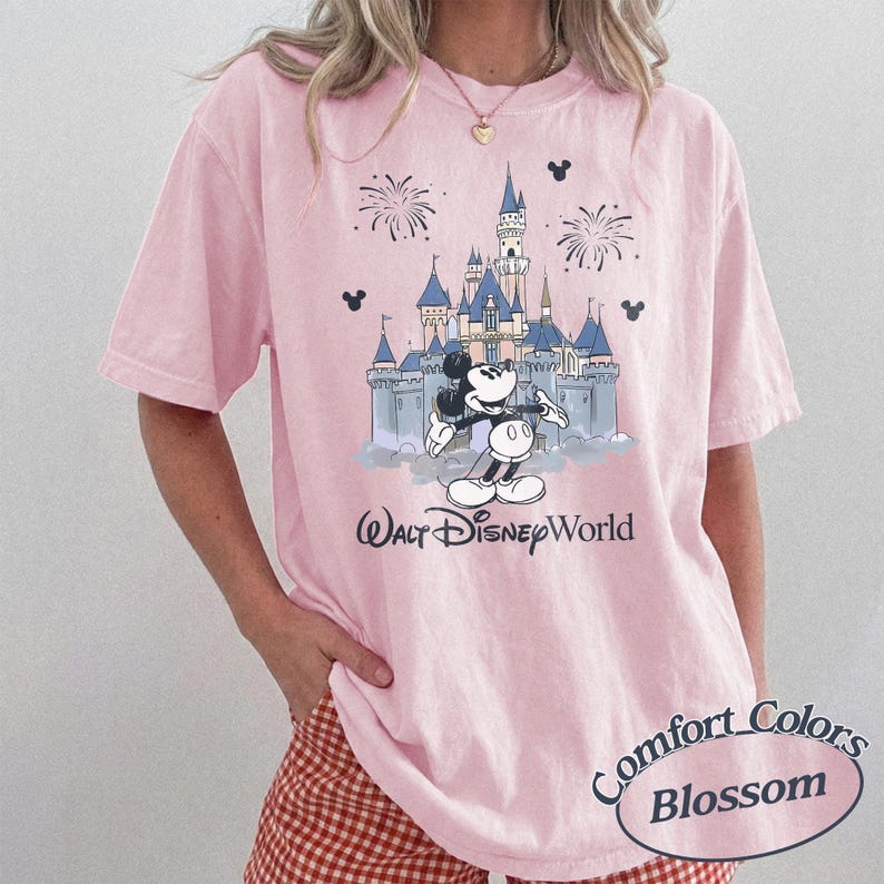 May include: A pale pink t-shirt with a graphic of Mickey Mouse and Cinderella's castle at Walt Disney World. The words "Walt Disney World" are printed below the graphic. The shirt also has the words "Comfort Colors Blossom" printed on the bottom right.