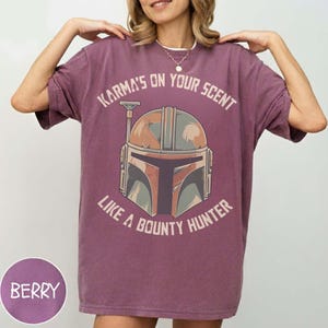 May include: Berry-coloured t-shirt featuring the text "Karma's on Your Scent Like a Bounty Hunter" and a Mandalorian helmet graphic. The shirt has a relaxed fit and short sleeves.