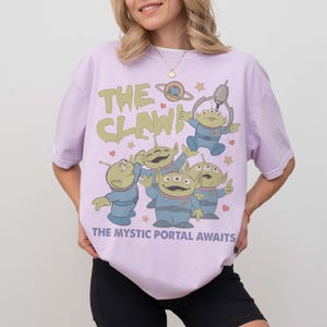 May include: A light purple t-shirt featuring a graphic of green aliens from the claw machine, with the text "THE CLAW!" and "THE MYSTIC PORTAL AWAITS". The aliens are wearing blue uniforms. The shirt has a relaxed fit.