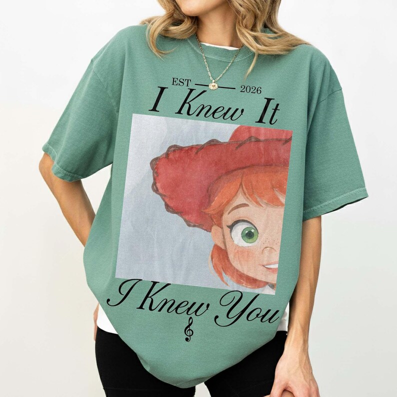May include: A sage green short-sleeve t-shirt featuring a graphic print of a cartoon character with red hair and a red hat. The text reads EST 2026, I Knew It, and I Knew You, with a treble clef symbol positioned below the final line of text.