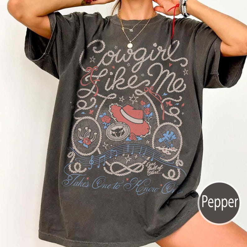 May include: A charcoal gray short-sleeve t-shirt featuring a graphic print with the text Cowgirl Like Me, Eyes full of Stars, and Takes One to Know One. The design includes a red cowboy hat, a horseshoe, rope motifs, musical notes, and stars.