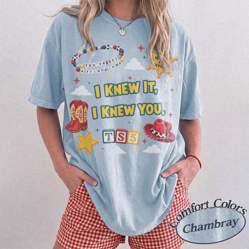 May include: A light blue short-sleeve t-shirt featuring a graphic print with the text I KNEW IT, I KNEW YOU, and TS5. The design includes a beaded bracelet, a yellow sheriff star, red cowboy boots, a red cowboy hat, and colorful alphabet blocks.