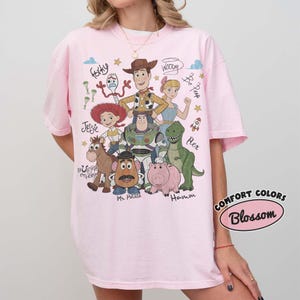 May include: A light pink t-shirt featuring a graphic print of Toy Story characters including Woody, Buzz Lightyear, Jessie, Rex, Hamm, Mr. Potato Head, Bullseye, Bo Peep, and Forky, with their names written in script and small star icons.