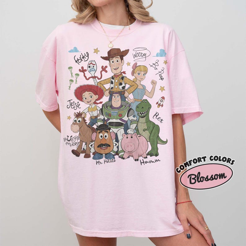May include: A light pink t-shirt featuring a graphic print of Toy Story characters including Woody, Buzz Lightyear, Jessie, Rex, Hamm, Mr. Potato Head, Bullseye, Bo Peep, and Forky, with their names written in script and small star icons.
