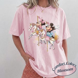 Op de afbeelding: Roze Comfort Colors T-shirt met een carrouselontwerp met Mickey en Minnie Mouse op witte paarden. Het ontwerp bevat pastelkleuren en de tekst "Comfort Colors Blossom" in een cirkel.