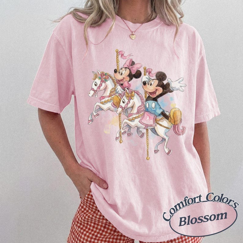 Op de afbeelding: Roze Comfort Colors T-shirt met een carrouselontwerp met Mickey en Minnie Mouse op witte paarden. Het ontwerp bevat pastelkleuren en de tekst "Comfort Colors Blossom" in een cirkel.