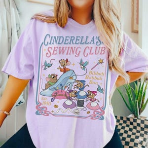 May include: A light purple t-shirt featuring a Cinderella's Sewing Club graphic. The design includes a blue glass slipper, mice, and the text "Bibbidi Bobbidi Boo." The shirt is a relaxed fit.