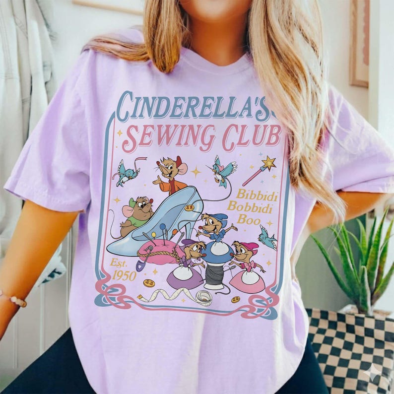 May include: A light purple t-shirt featuring a Cinderella's Sewing Club graphic. The design includes a blue glass slipper, mice, and the text "Bibbidi Bobbidi Boo." The shirt is a relaxed fit.