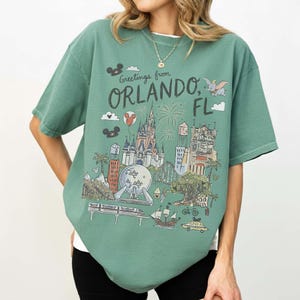 K&ouml;nnte beinhalten: Gr&uuml;nes T-Shirt mit einer Grafik von Orlando, Florida, mit einem Schloss, Feuerwerk und einer Einschienenbahn. Der Text "Greetings from Orlando, FL" ist auf der Vorderseite aufgedruckt. Das Shirt hat kurze &Auml;rmel und eine lockere Passform.