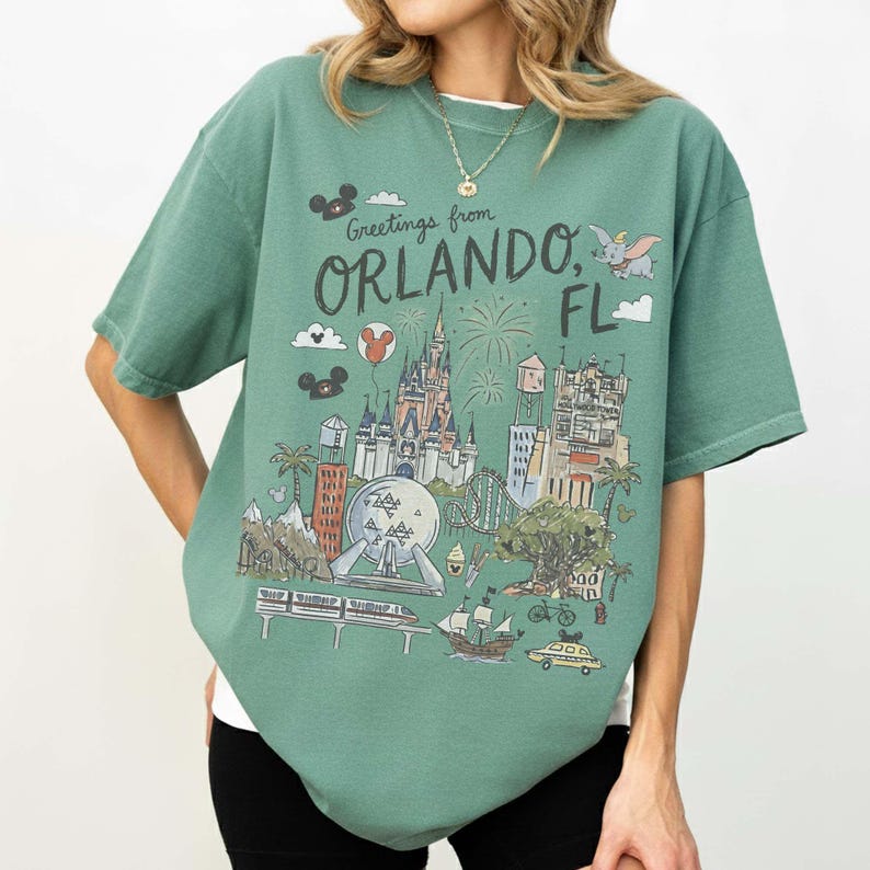 K&ouml;nnte beinhalten: Gr&uuml;nes T-Shirt mit einer Grafik von Orlando, Florida, mit einem Schloss, Feuerwerk und einer Einschienenbahn. Der Text "Greetings from Orlando, FL" ist auf der Vorderseite aufgedruckt. Das Shirt hat kurze &Auml;rmel und eine lockere Passform.