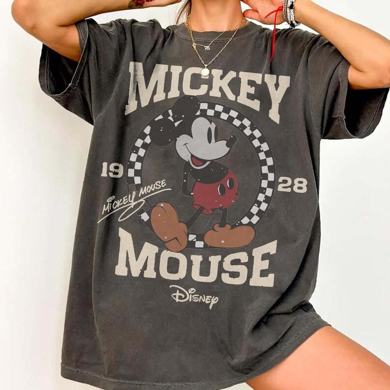 May include: Dark gray t-shirt featuring Mickey Mouse. The design includes the words "Mickey Mouse" in large letters, the years "1928" and the Disney logo. The cartoon character is in the center of a checkered circle.
