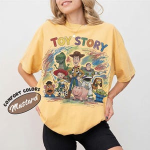Pu&ograve; includere: Una t-shirt giallo senape con una stampa grafica colorata dei personaggi di Toy Story, tra cui Woody, Buzz Lightyear, Jessie, Rex, Hamm, Slinky Dog, Mr. Potato Head e Alien. Il testo TOY STORY &egrave; stampato in un carattere multicolore sopra.