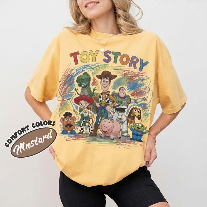 Pu&ograve; includere: Una t-shirt giallo senape con una stampa grafica colorata dei personaggi di Toy Story, tra cui Woody, Buzz Lightyear, Jessie, Rex, Hamm, Slinky Dog, Mr. Potato Head e Alien. Il testo TOY STORY &egrave; stampato in un carattere multicolore sopra.