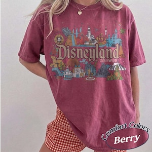 May include: Berry-colored t-shirt with a Disneyland Resort graphic. The design features various park icons and the Disneyland Resort logo. The shirt is a Comfort Colors brand.
