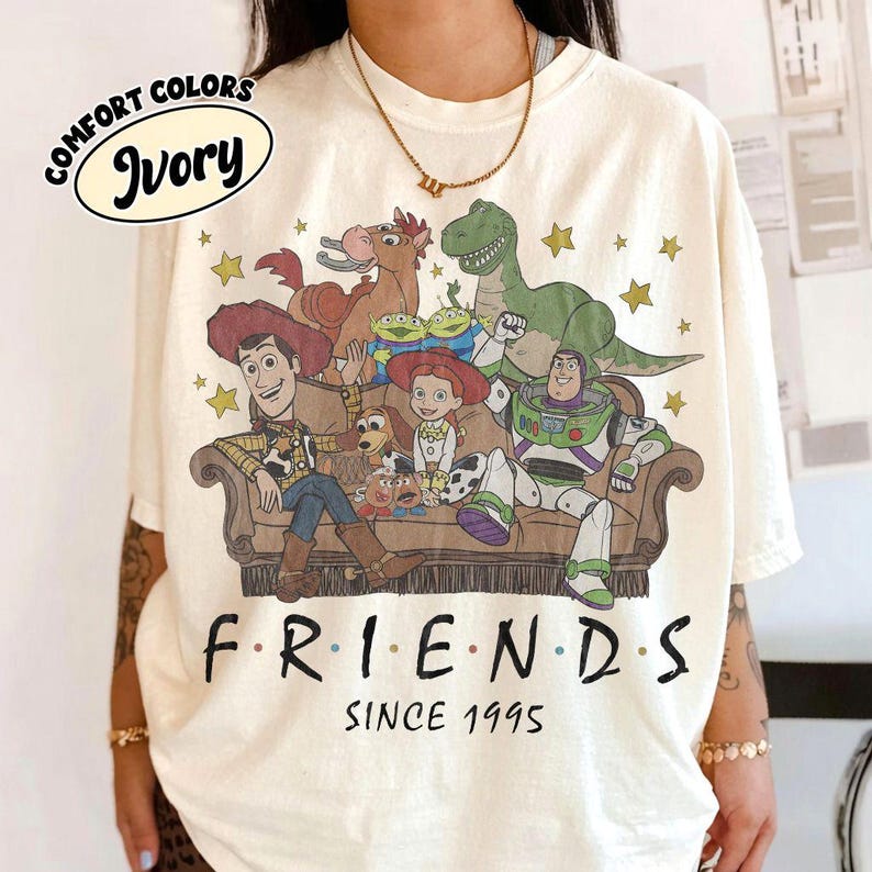 May include: An ivory-colored t-shirt featuring a graphic print of Toy Story characters sitting on a couch, including Woody, Buzz Lightyear, Jessie, Bullseye, Rex, Slinky Dog, Mr. Potato Head, and Aliens. The text reads F R I E N D S SINCE 1995.