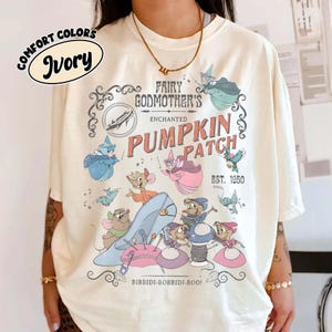 May include: Ivory-colored t-shirt featuring a whimsical design with the text "FAIRY GODMOTHER'S ENCHANTED PUMPKIN PATCH EST. 1950". The graphic includes Cinderella-themed illustrations and the phrase "BIBBIDI-BOBBIDI-BOO!".