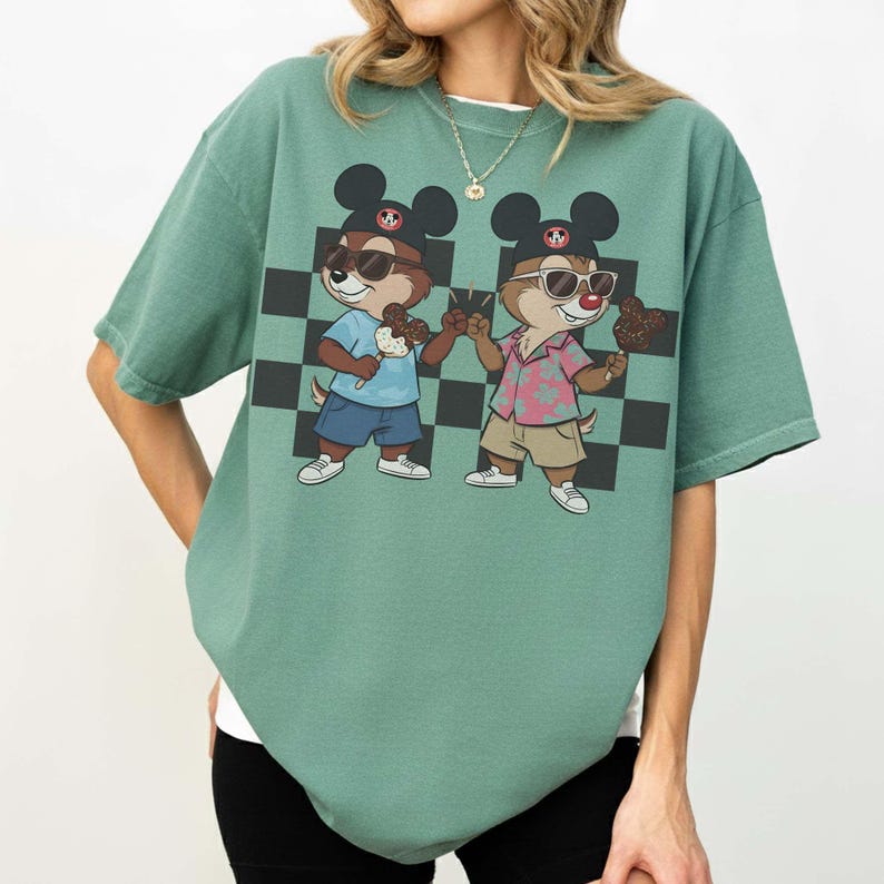 May include: A green t-shirt featuring a cartoon graphic of two chipmunks wearing sunglasses and Mickey Mouse ears. They are holding ice cream treats in front of a black and white checkered background. The shirt has short sleeves.