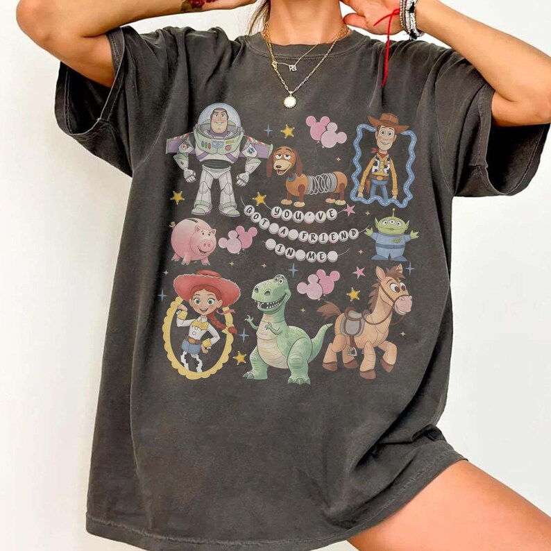 K&ouml;nnte beinhalten: Dunkelgraues T-Shirt mit einer farbenfrohen Grafik von Charakteren aus einem beliebten Zeichentrickfilm. Das Design umfasst Buzz Lightyear, Woody, Jessie, Rex und andere, mit dem Satz "You've Got a Friend in Me."