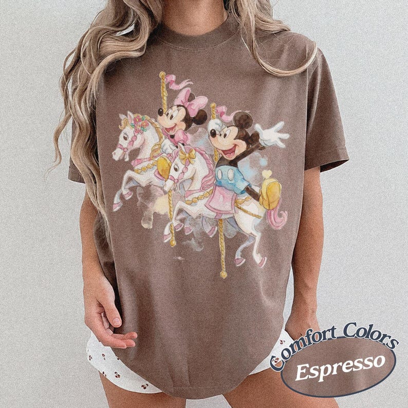 Op de afbeelding: Espresso-kleurig T-shirt met een carrouselsc&egrave;ne met Mickey en Minnie Mouse op witte paarden. Het ontwerp bevat roze en gele accenten. Het shirt is voorzien van het opschrift "Comfort Colors" en "Espresso" in een cursief lettertype.