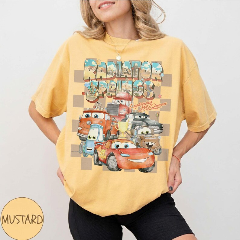 Peut inclure: T-shirt moutarde avec un motif des personnages du film Cars, dont Flash McQueen, avec le texte "Radiator Springs". Le motif est plac&eacute; sur un fond &agrave; carreaux.