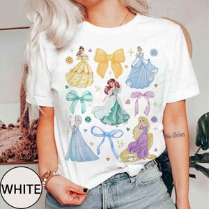 May include: White t-shirt featuring watercolor illustrations of princesses and bows. The design includes images of princesses in yellow, blue, and green gowns, along with various colored bows and floral accents. The word "WHITE" is printed on the bottom left.