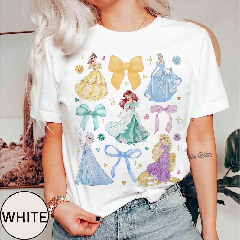 May include: White t-shirt featuring watercolor illustrations of princesses and bows. The design includes images of princesses in yellow, blue, and green gowns, along with various colored bows and floral accents. The word "WHITE" is printed on the bottom left.