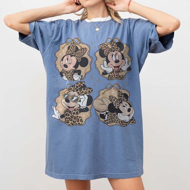 May include: A blue t-shirt dress featuring four Minnie Mouse designs. Each design shows Minnie in a leopard print outfit, set within a tan and gold frame. The dress has short sleeves and a relaxed fit.