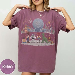 Puede incluir: Camiseta de color bayas con un gr&aacute;fico de un parque tem&aacute;tico, fuegos artificiales y personajes de dibujos animados. El dise&ntilde;o incluye una gran esfera, edificios y figuras animadas. La camiseta tiene un corte holgado.