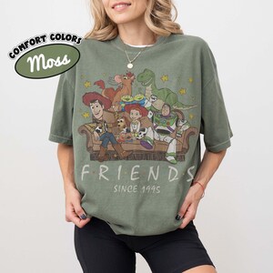 May include: A moss green short-sleeve t-shirt featuring a graphic print of Toy Story characters sitting on a couch with the text "FRIENDS SINCE 1995" below the illustration. The shirt has a crew neckline and a relaxed, oversized fit.