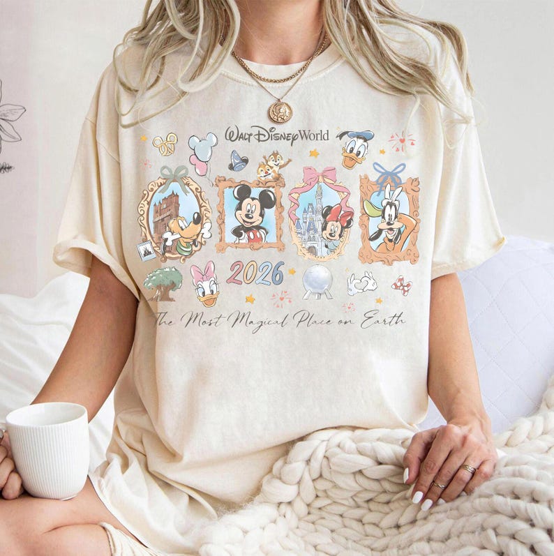 May include: Cream-colored t-shirt featuring a colorful graphic of Disney characters in framed portraits. The design includes Mickey Mouse, Minnie Mouse, Donald Duck, and Goofy, with the text "Walt Disney World" and "2026". The shirt also has the phrase "The Most Magical Place on Earth."