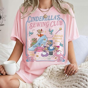 May include: Pink t-shirt with a graphic featuring Cinderella's Sewing Club. The design includes a blue glass slipper, mice, birds, and the text "Bibbidi Bobbidi Boo." The shirt is dated "Est. 1950."