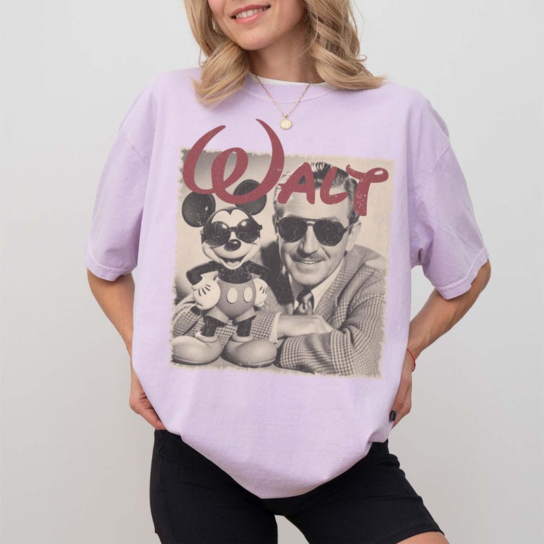 Pu&ograve; includere: Una t-shirt a maniche corte color viola chiaro con una stampa grafica di Topolino e Walt Disney, entrambi con occhiali da sole. Il testo "WALT" &egrave; stampato in un carattere rosso dall'aspetto vissuto sopra l'immagine in bianco e nero.