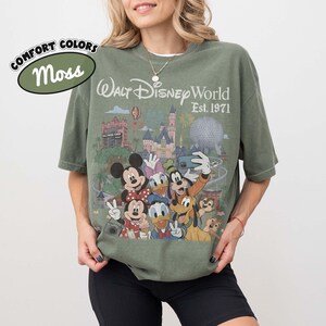 May include: A moss green short-sleeve t-shirt featuring a graphic print of Walt Disney World Est. 1971 with Mickey Mouse, Minnie Mouse, Donald Duck, Daisy Duck, Goofy, Pluto, Chip, and Dale taking a selfie in front of the park landmarks.
