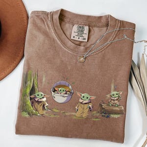 May include: A brown short-sleeve t-shirt featuring a graphic print of four small green creatures with large ears. One creature stands by a tree, one is in a floating pod, one stands on the ground, and one sits on a log. The shirt has a Comfort Colors label.