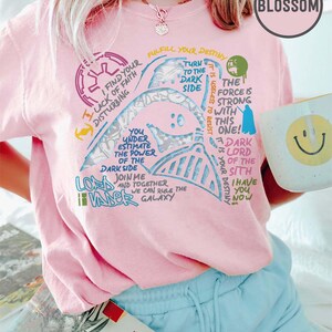 May include: Pale pink t-shirt with a Darth Vader graphic and Star Wars quotes. The design includes phrases such as "Turn to the Dark Side" and "The Force is Strong." A white mug with a smiley face is held.