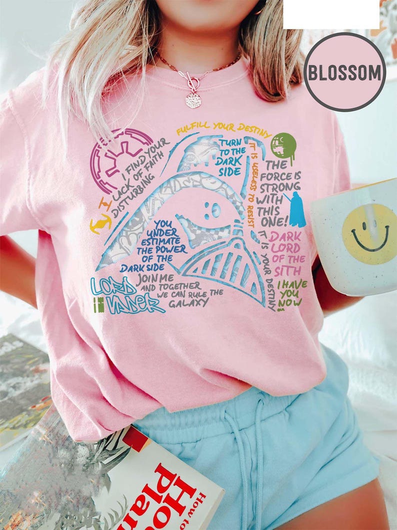 May include: Pale pink t-shirt with a Darth Vader graphic and Star Wars quotes. The design includes phrases such as "Turn to the Dark Side" and "The Force is Strong." A white mug with a smiley face is held.