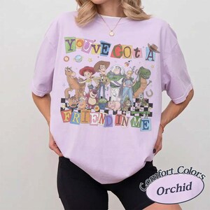 May include: A light purple short-sleeve t-shirt featuring a colorful graphic of various animated characters from the Toy Story franchise. The text "YOU'VE GOT A FRIEND IN ME" is printed in multi-colored block letters above and below the characters.