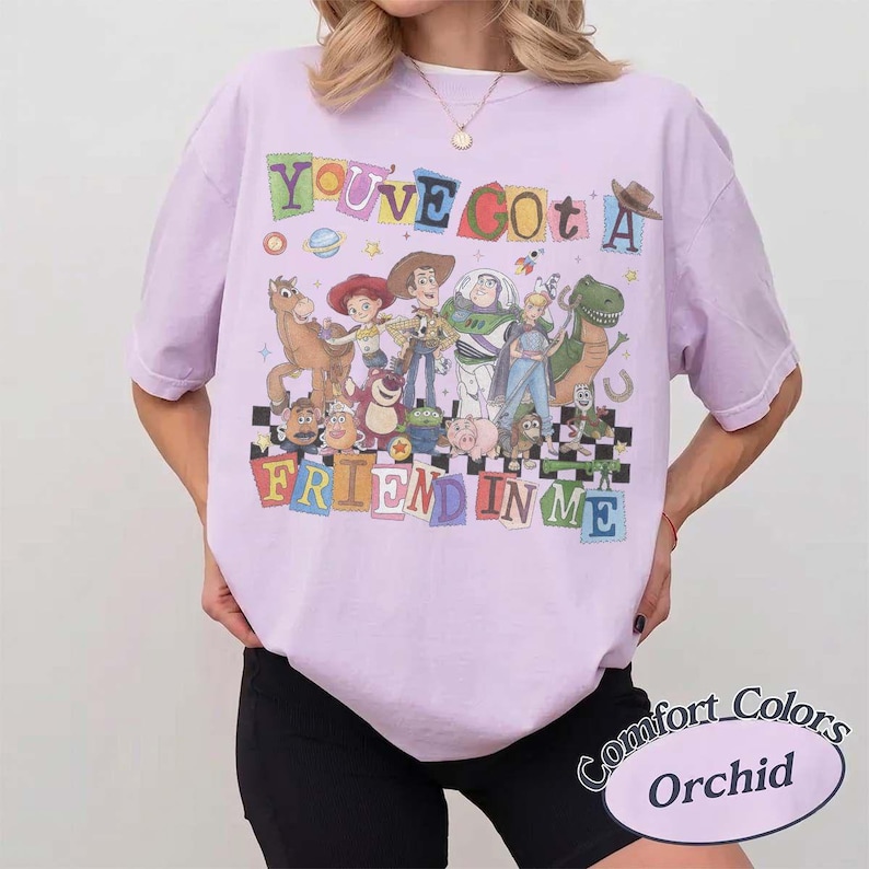 May include: A light purple short-sleeve t-shirt featuring a colorful graphic of various animated characters from the Toy Story franchise. The text "YOU'VE GOT A FRIEND IN ME" is printed in multi-colored block letters above and below the characters.