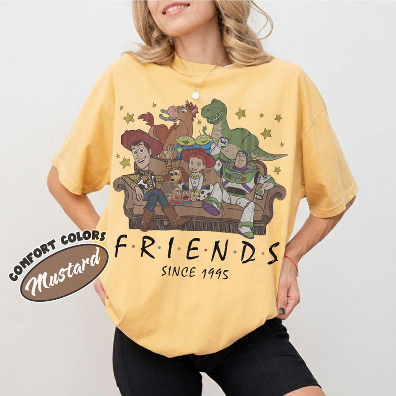 May include: A mustard yellow short-sleeve t-shirt featuring a graphic of Toy Story characters sitting on a couch. The text reads FRIENDS SINCE 1995. The shirt has a crew neckline and a relaxed fit. A small oval label indicates the color is mustard.