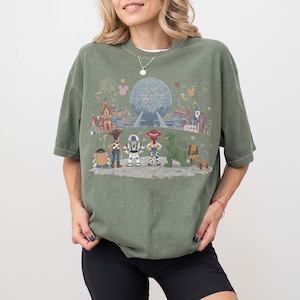 Puede incluir: Camiseta verde oliva con un gr&aacute;fico colorido de personajes de Disney y una escena de parque. El dise&ntilde;o incluye personajes como Woody, Buzz Lightyear y Rex, con una gran esfera y edificios en el fondo. La camiseta tiene mangas cortas y un corte holgado.