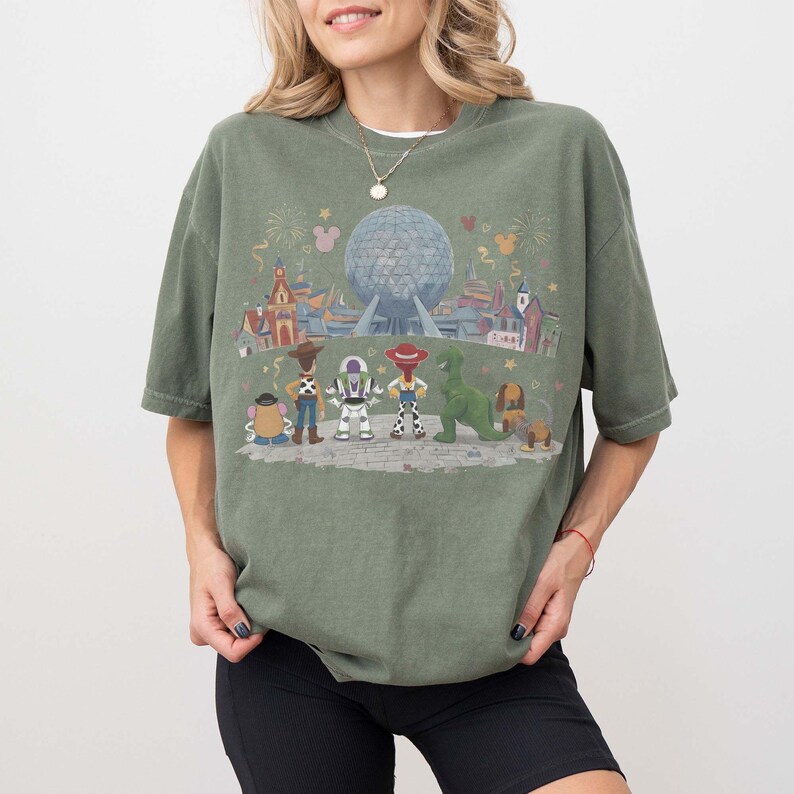Puede incluir: Camiseta verde oliva con un gr&aacute;fico colorido de personajes de Disney y una escena de parque. El dise&ntilde;o incluye personajes como Woody, Buzz Lightyear y Rex, con una gran esfera y edificios en el fondo. La camiseta tiene mangas cortas y un corte holgado.