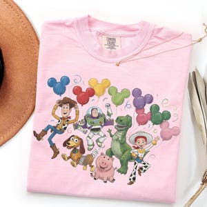 May include: A light pink t-shirt featuring a graphic print of Toy Story characters including Woody, Buzz Lightyear, Rex, Slinky Dog, Hamm, and Jessie. The characters are surrounded by colorful Mickey Mouse-shaped balloons and confetti.