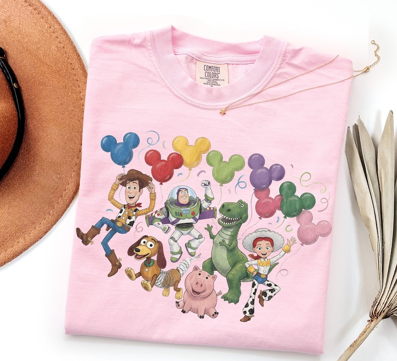 May include: A light pink t-shirt featuring a graphic print of Toy Story characters including Woody, Buzz Lightyear, Rex, Slinky Dog, Hamm, and Jessie. The characters are surrounded by colorful Mickey Mouse-shaped balloons and confetti.