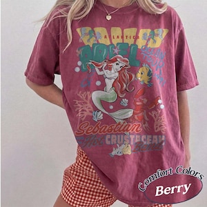 May include: Berry-colored t-shirt featuring a graphic of Ariel from The Little Mermaid with the text "A Lastic" and "Scan Your Tail". The shirt also features the characters Sebastian and Flounder.