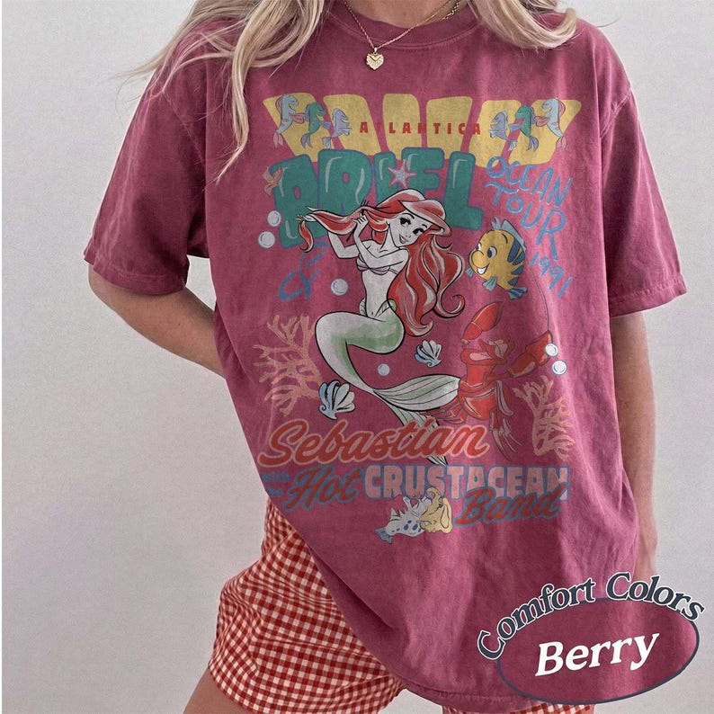 May include: Berry-colored t-shirt featuring a graphic of Ariel from The Little Mermaid with the text "A Lastic" and "Scan Your Tail". The shirt also features the characters Sebastian and Flounder.