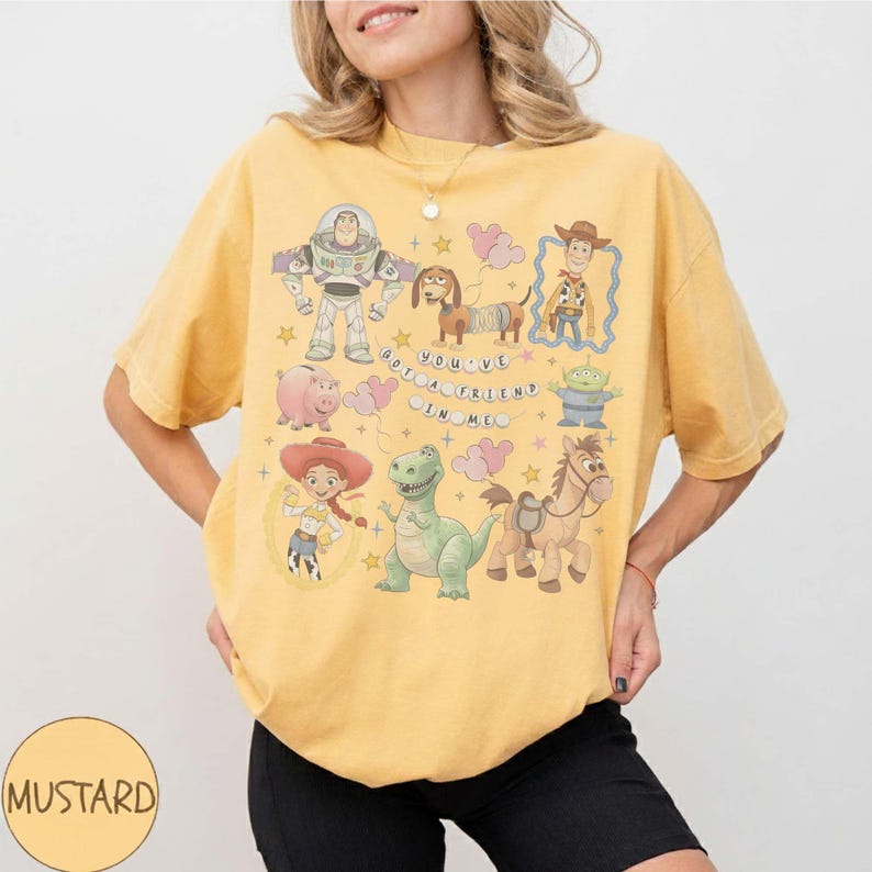 K&ouml;nnte beinhalten: Senffarbenes T-Shirt mit einer Grafik von Charakteren aus dem Animationsfilm "Toy Story". Das Design umfasst Buzz Lightyear, Woody, Jessie und andere Charaktere mit dem Text "You've Got a Friend in Me".
